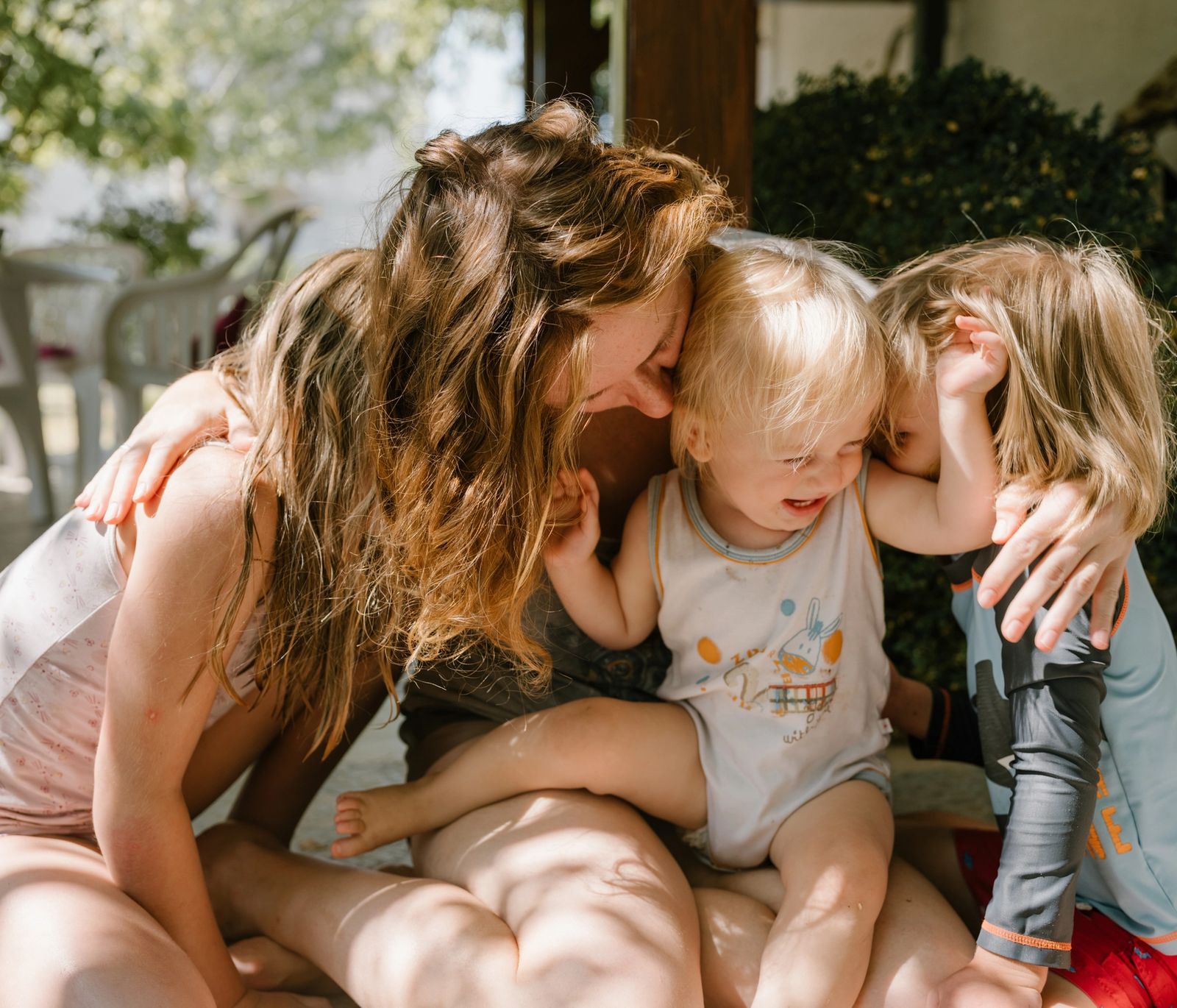 A woman sits on the ground with two young children draped over her, all of them laughing.