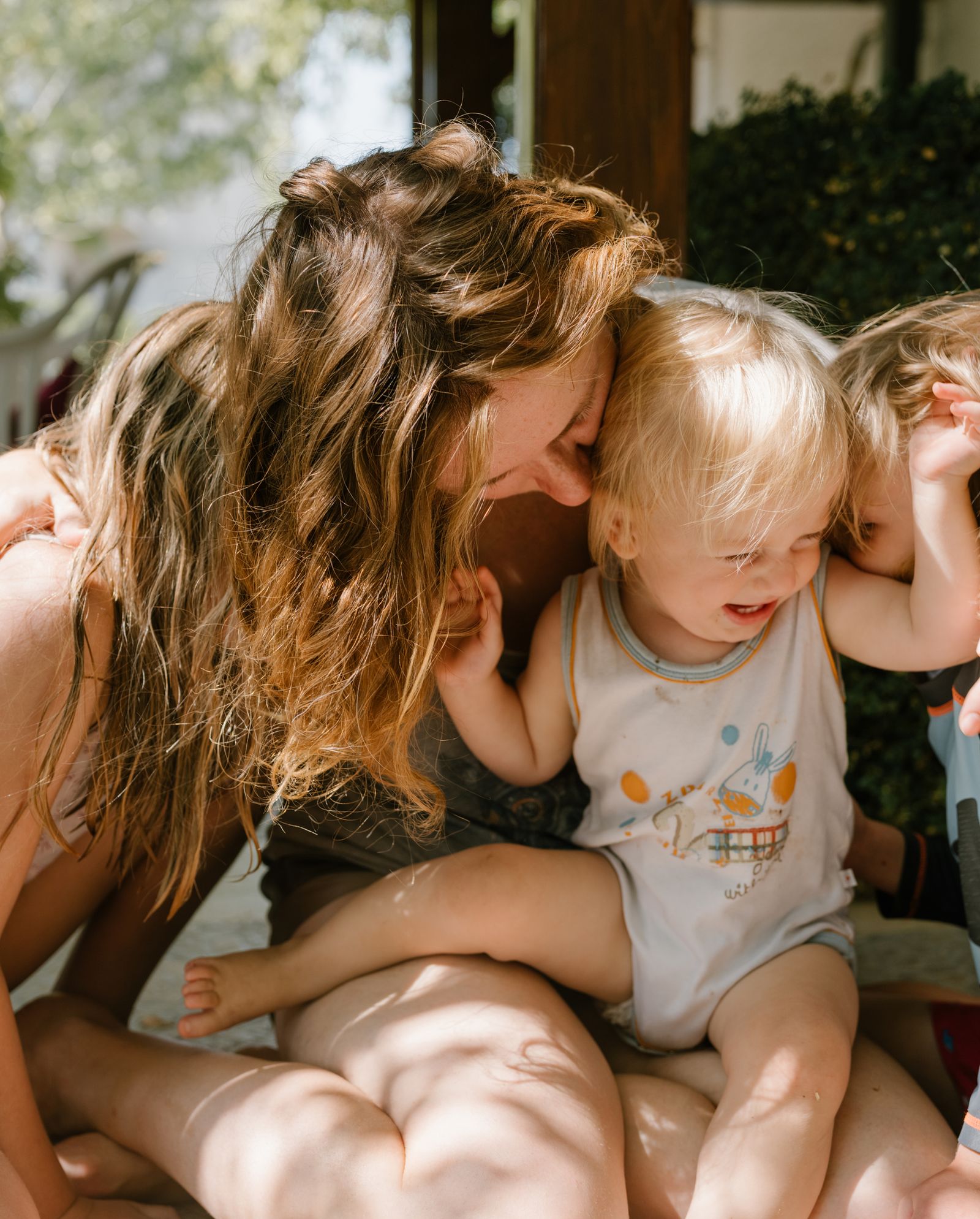 A woman sits on the ground with two young children draped over her, all of them laughing.
