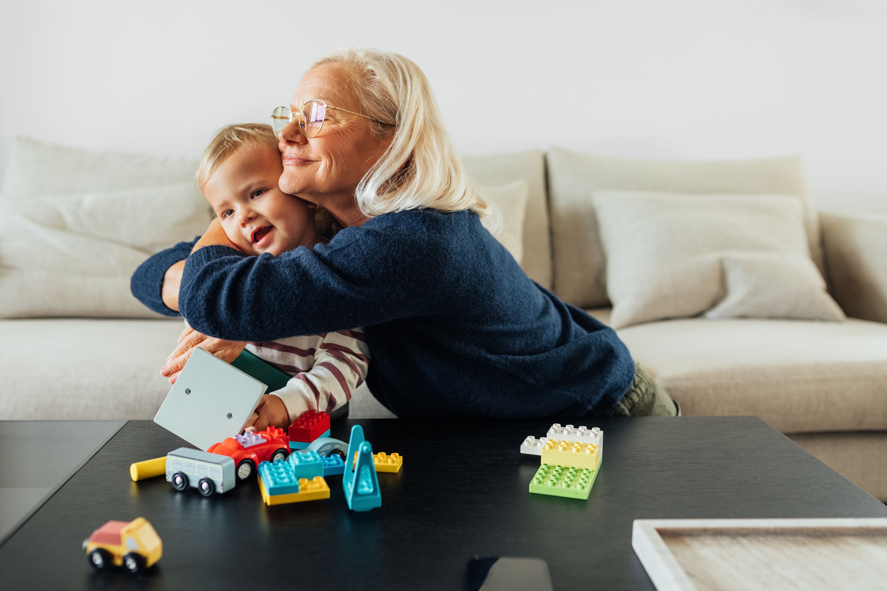 An older woman hugs a small child on a sofa, building blocks scattered on the table in front of them.