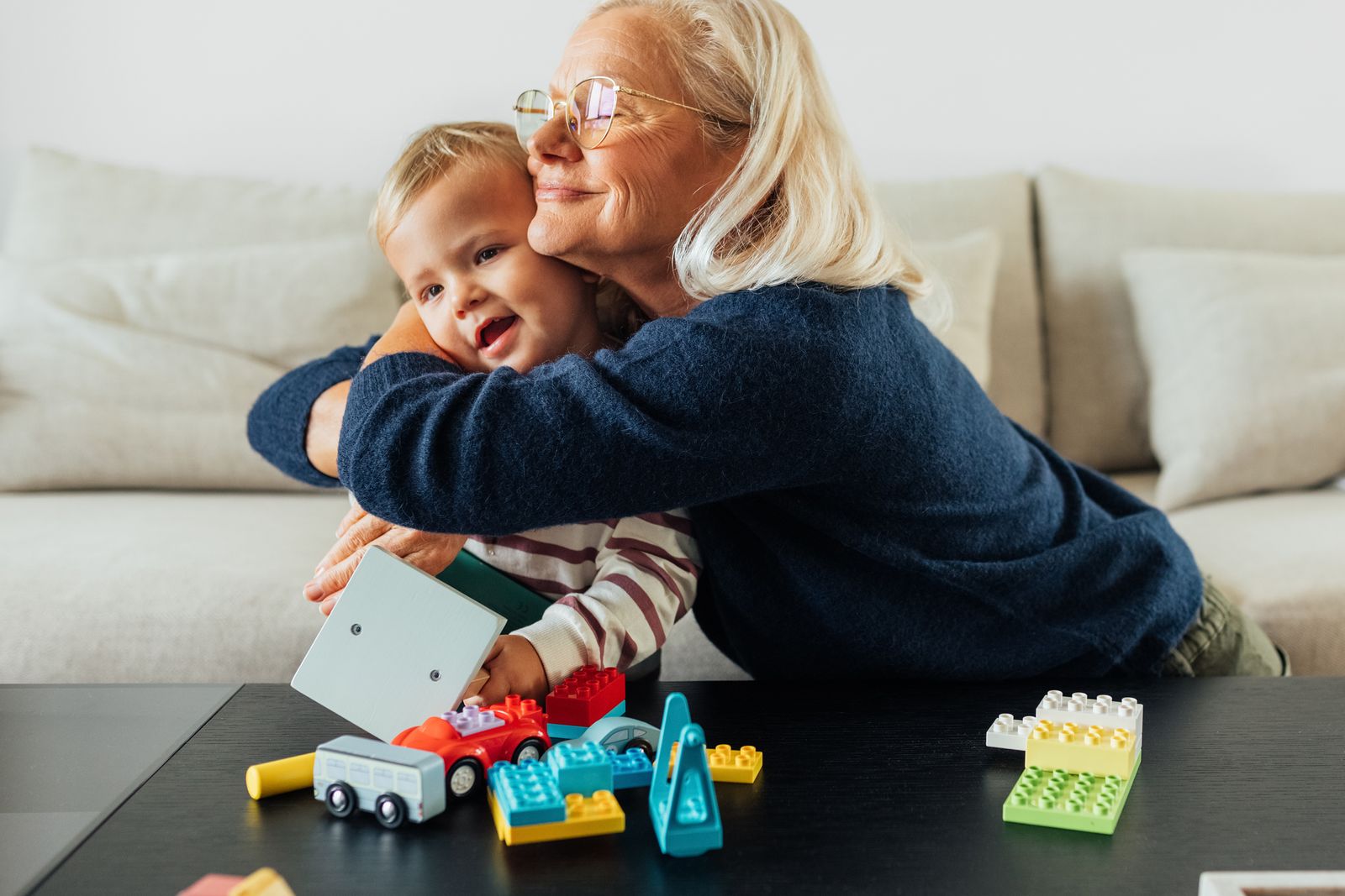 An older woman hugs a small child on a sofa, building blocks scattered on the table in front of them.