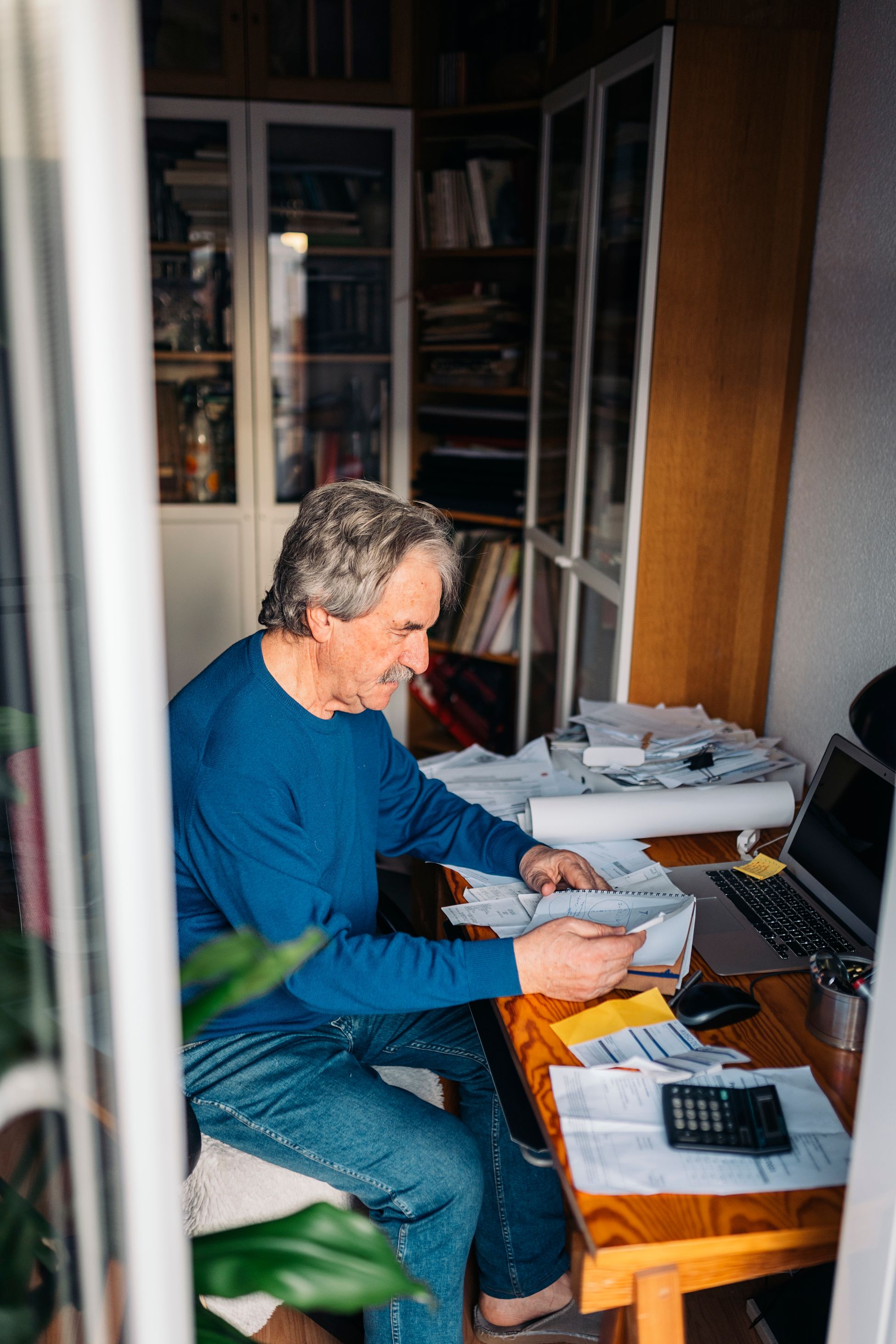 An older man sits at a cluttered home desk sorting through a stack of receipts beside a laptop and calculator.
