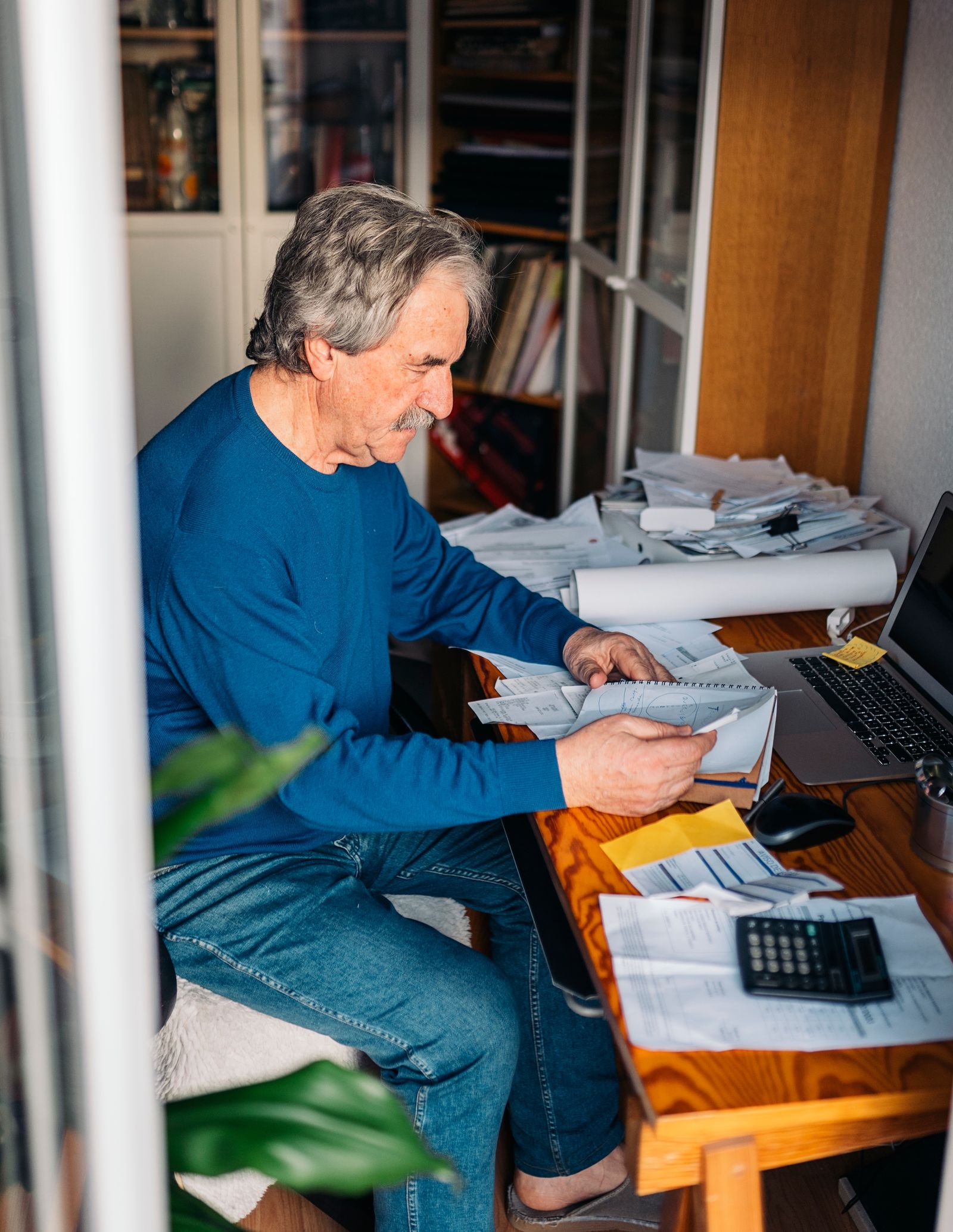 An older man sits at a cluttered home desk sorting through a stack of receipts beside a laptop and calculator.