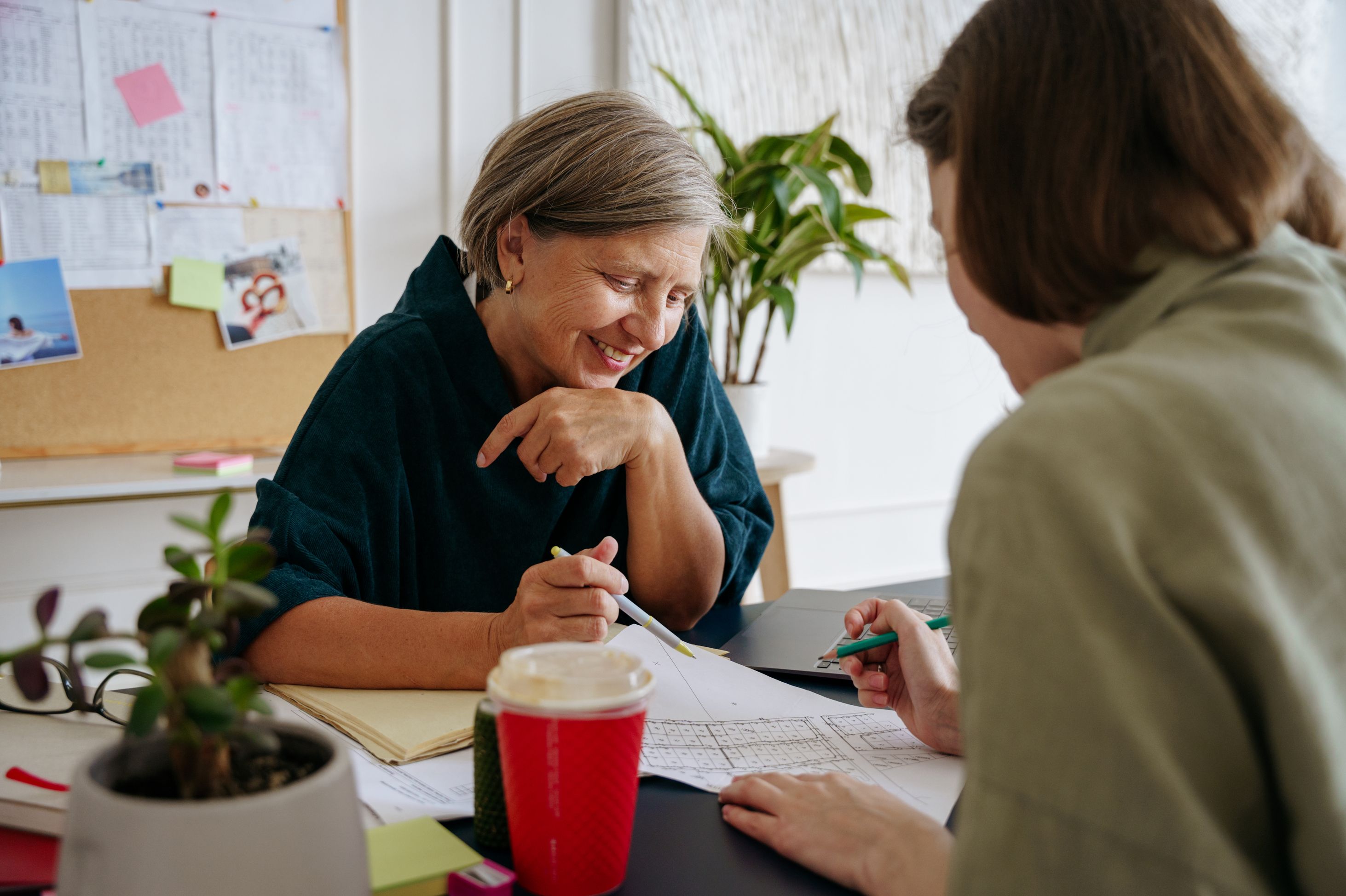Two women sit across a table, smiling as they go over documents and drawings together.