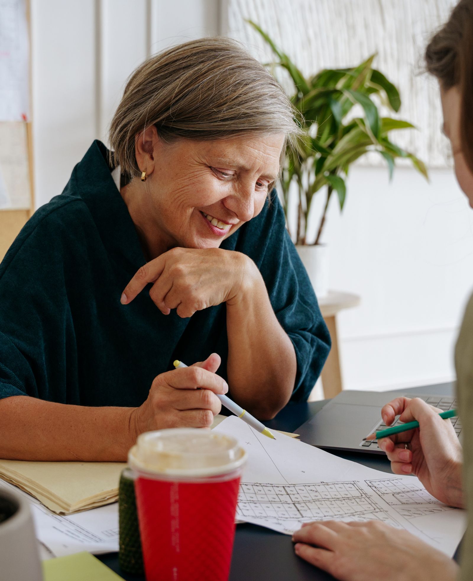 Two women sit across a table, smiling as they go over documents and drawings together.