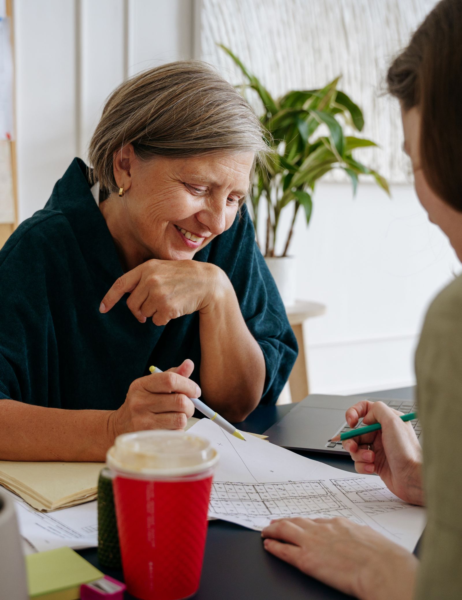 Two women sit across a table, smiling as they go over documents and drawings together.