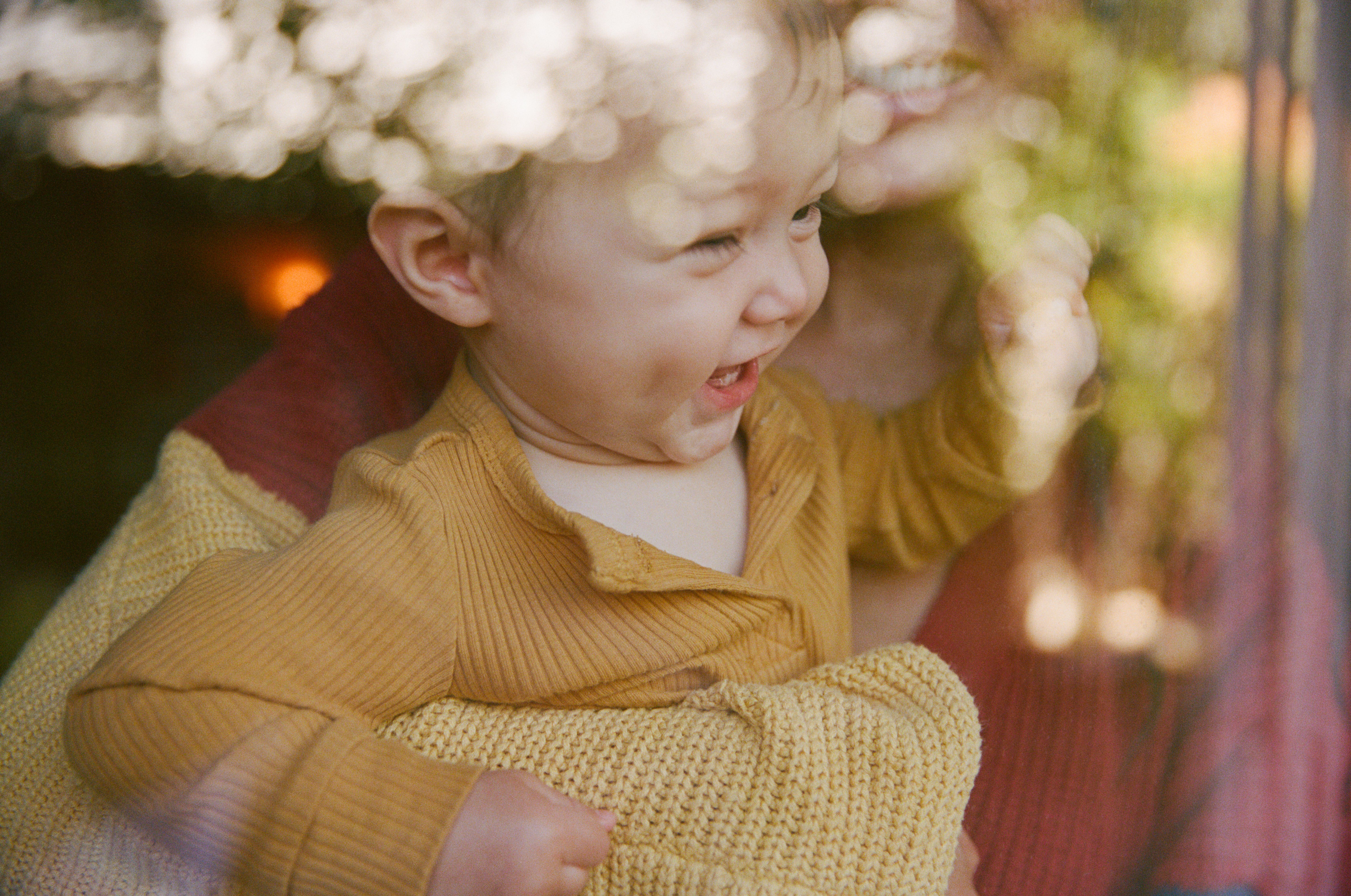 A baby in a yellow knit cardigan laughs while being held outdoors in dappled light.
