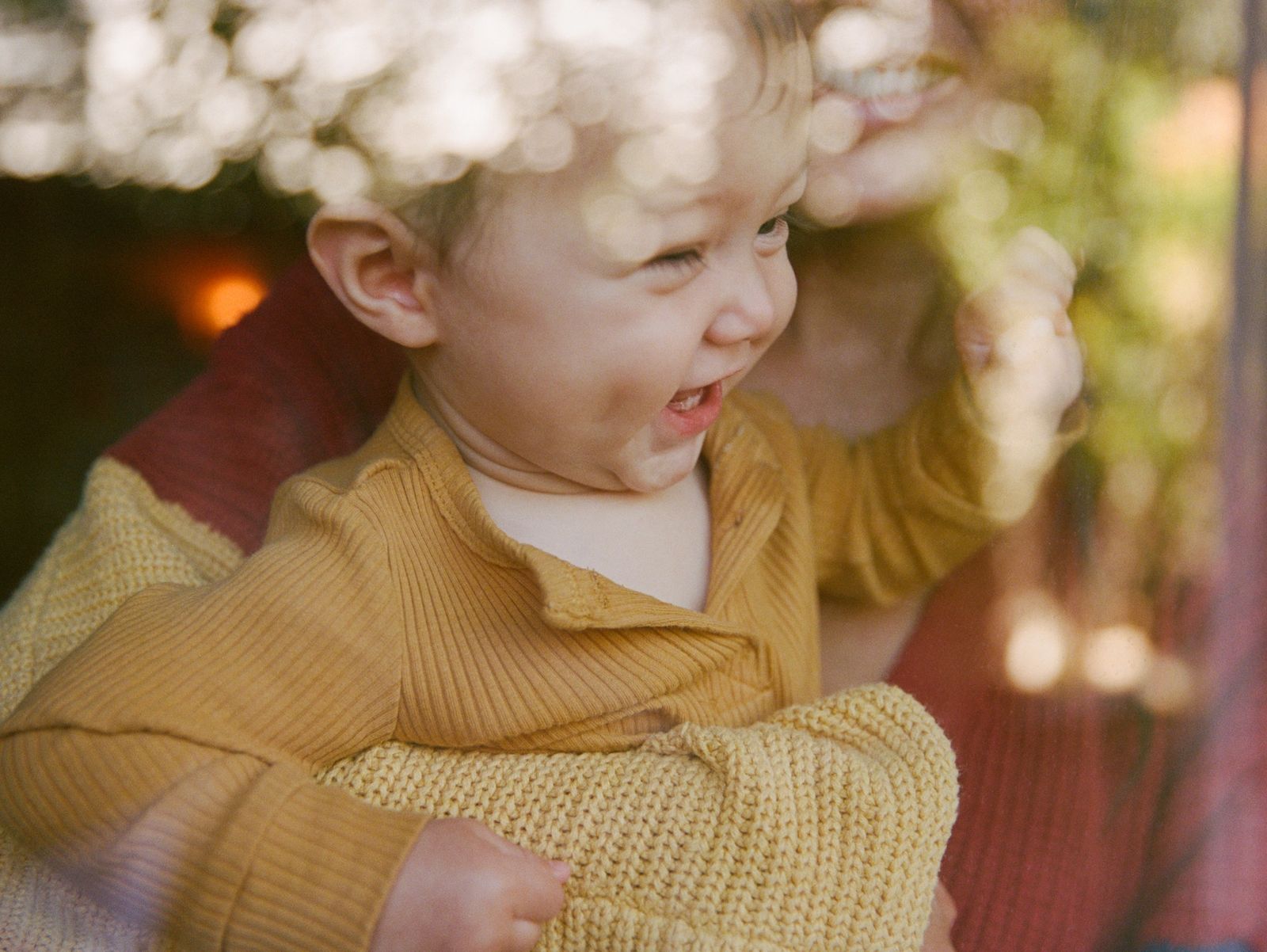 A baby in a yellow knit cardigan laughs while being held outdoors in dappled light.