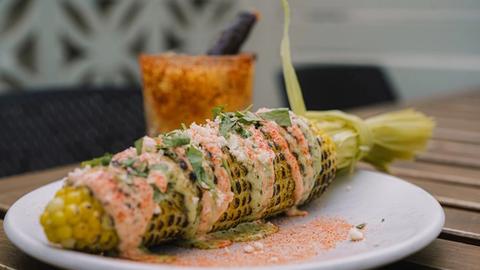 a plate of charred corn on the cob with cilantro and chipotle and cilantro aioli. in the background is a mango margarita with tajin rim.