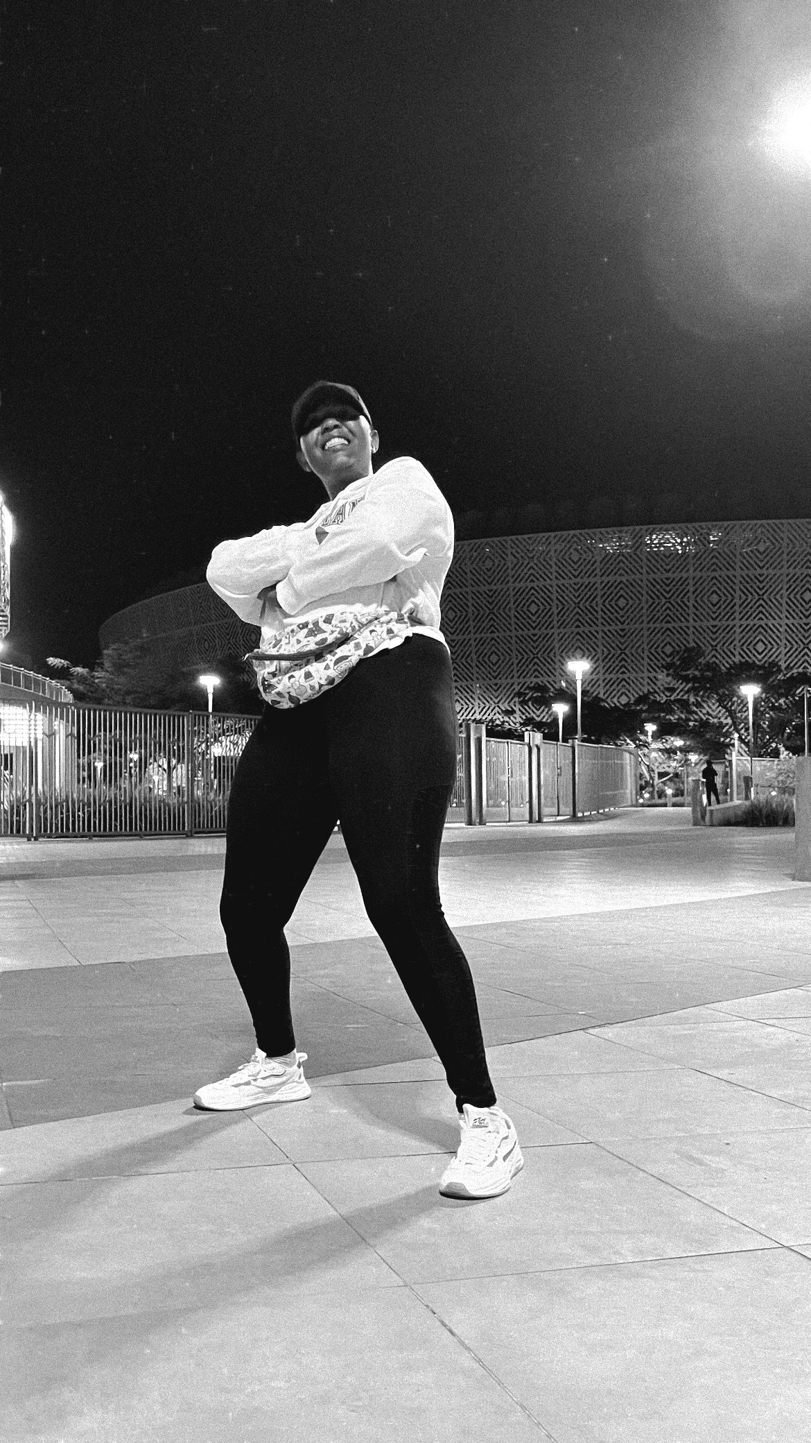 Natasha Umrungi in black and white outside a floodlit arena at night, arms folded, in a cap and white sweatshirt, laughing.