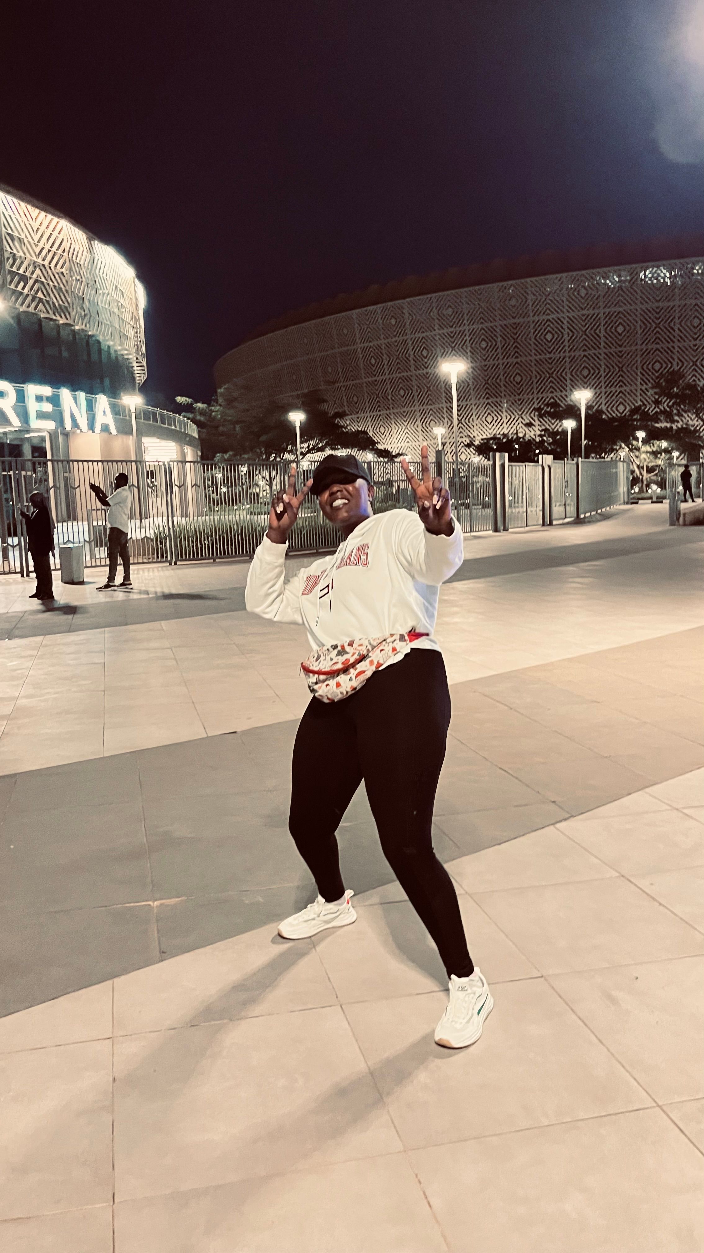 Natasha Umrungi outside a lit arena at night, both hands raised in peace signs, grinning, in a cap and white sweatshirt.