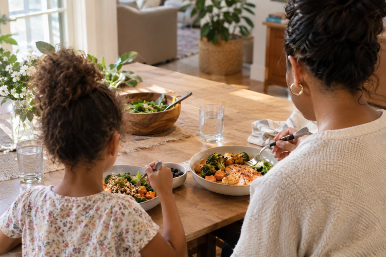 A woman and a young girl eating at a sunlit wooden table, with bowls of roasted salmon, broccoli, and sweet potato in front of them