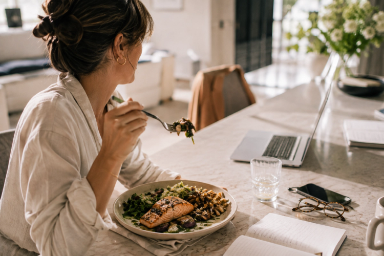 A woman eats a plate of seared salmon, greens, and roasted vegetables at a marble kitchen counter next to an open laptop, a mug, and glasses.