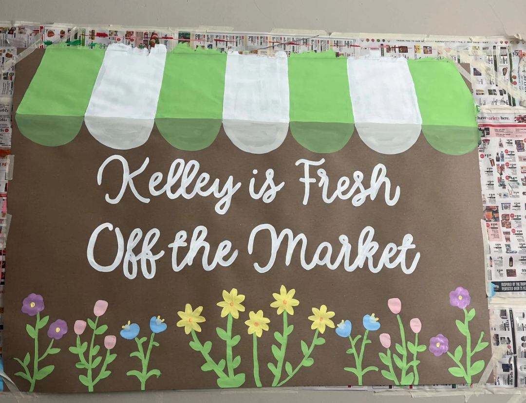 Kraft paper banner reading "Kelley is Fresh Off the Market" in white script, under a green and white striped market awning, with a row of hand-painted wildflowers along the bottom.
