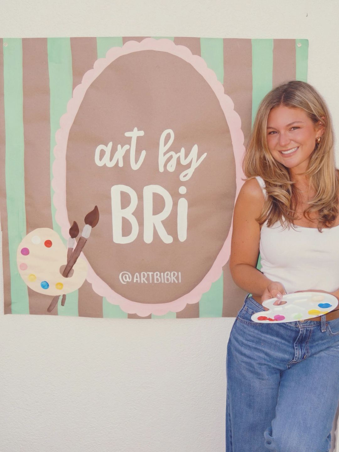 Bri smiling beside her hand-painted "art by Bri" shop banner, with finished banners rolled and wrapped ready to go.