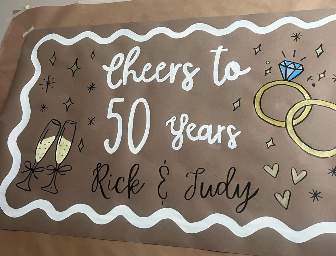 Anniversary banner reading "Cheers to 50 Years, Rick & Judy" inside a white wavy border, with painted champagne flutes, wedding rings and hearts.