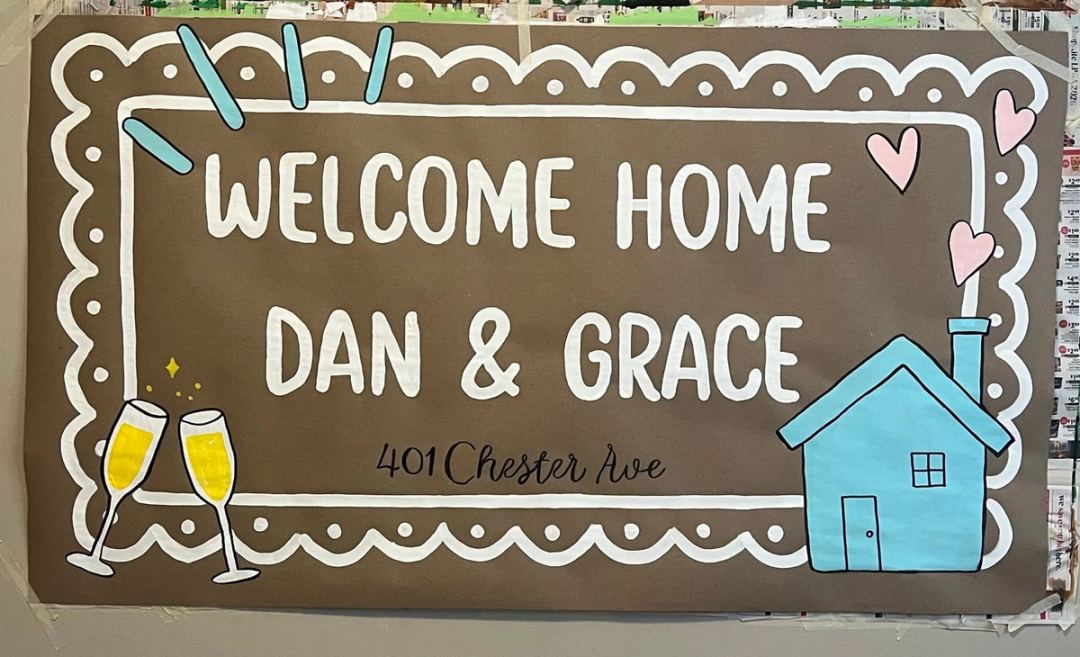 Housewarming banner reading "Welcome Home Dan & Grace, 401 Chester Ave" inside a scalloped white border, with a painted blue house and champagne glasses.