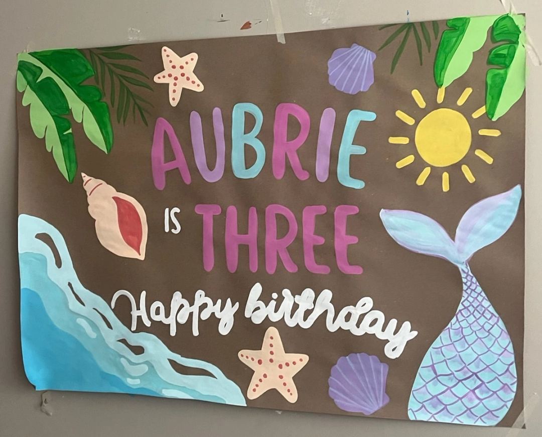 Under-the-sea birthday banner reading "Aubrie is Three, Happy Birthday" with a painted mermaid tail, starfish, seashells, palm leaves and a sun.