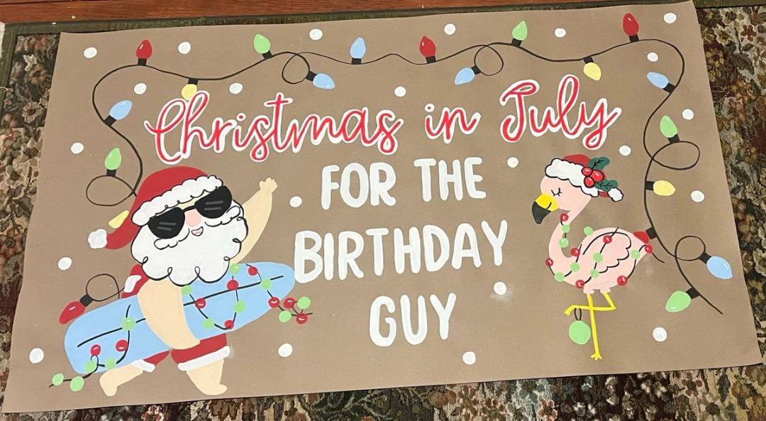 Banner reading "Christmas in July for the Birthday Guy" with a painted Santa in sunglasses and swim trunks, a flamingo in a Santa hat, and a string of Christmas lights.
