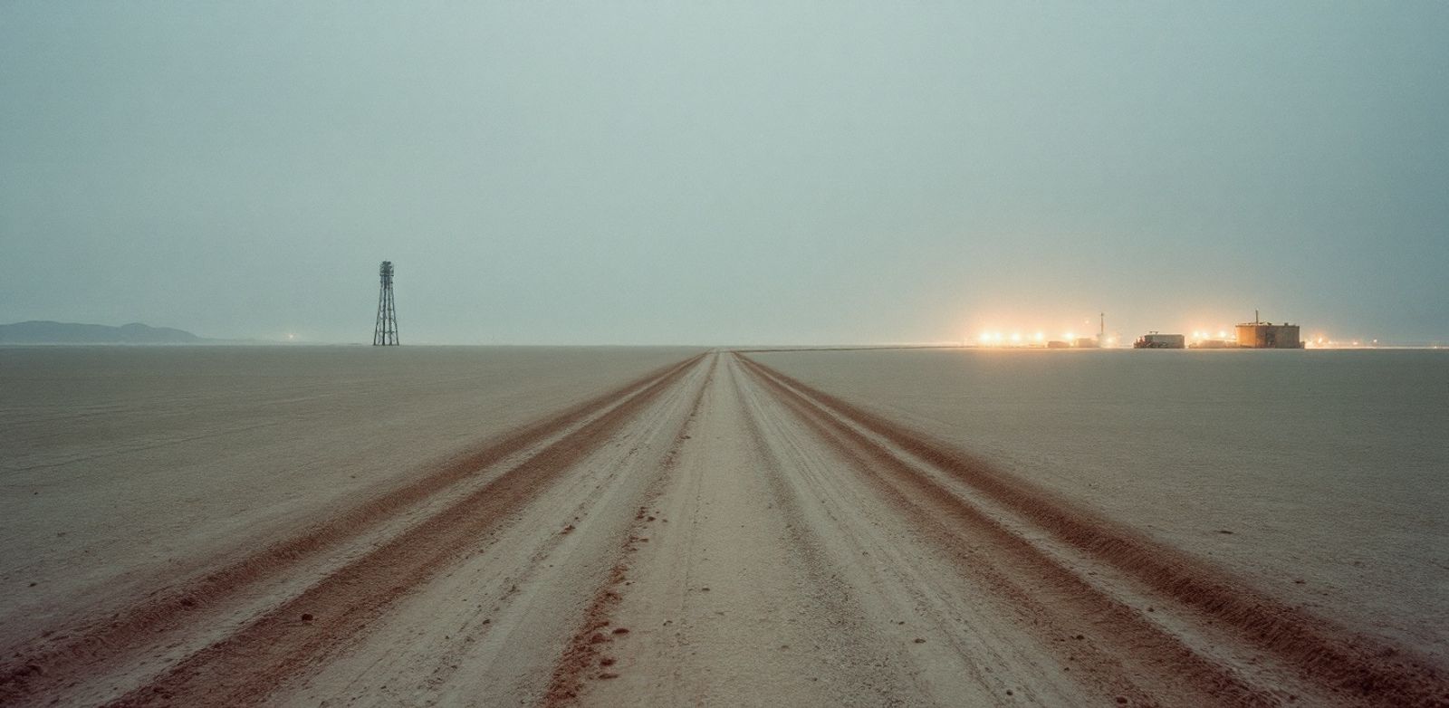 Dry salt flat with two paths leading toward a tower and distant warehouse lights