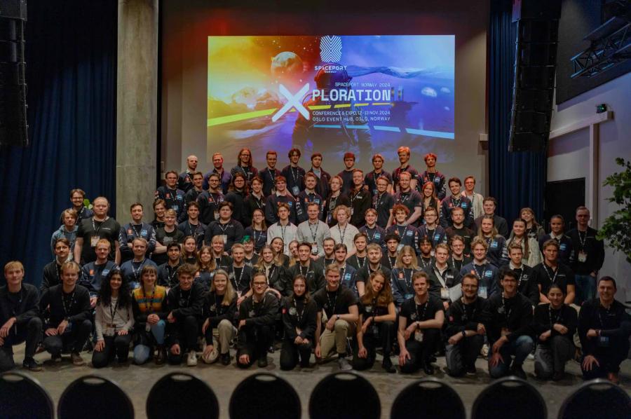 A large group of NORSTEC members gathered in front of a colorful presentation screen during the Xploration event, posing together for a group photo indoors.