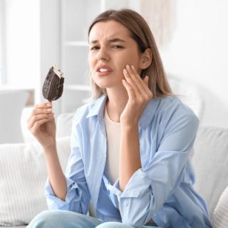 Image of a woman at her home eating an ice cream, she is holding her face due to dental issues.