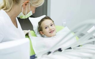 A dentist in scrubs stands beside a young child seated in a dental chair, with the young girl smiling at the dentist. The bright and inviting dental clinic exudes a welcoming atmosphere, reflected in their cheerful expressions.