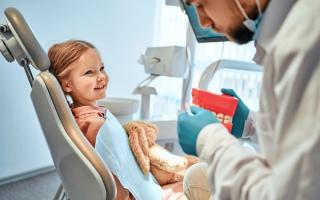 A dentist wearing scrubs smiles warmly while standing next to a young child in a dental chair, both looking at the camera with cheerful expressions in a bright, welcoming dental clinic.