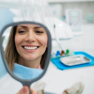 Image of a woman holding a small mirror looking at her teeth, she is in a dental clinic under going an appointment.