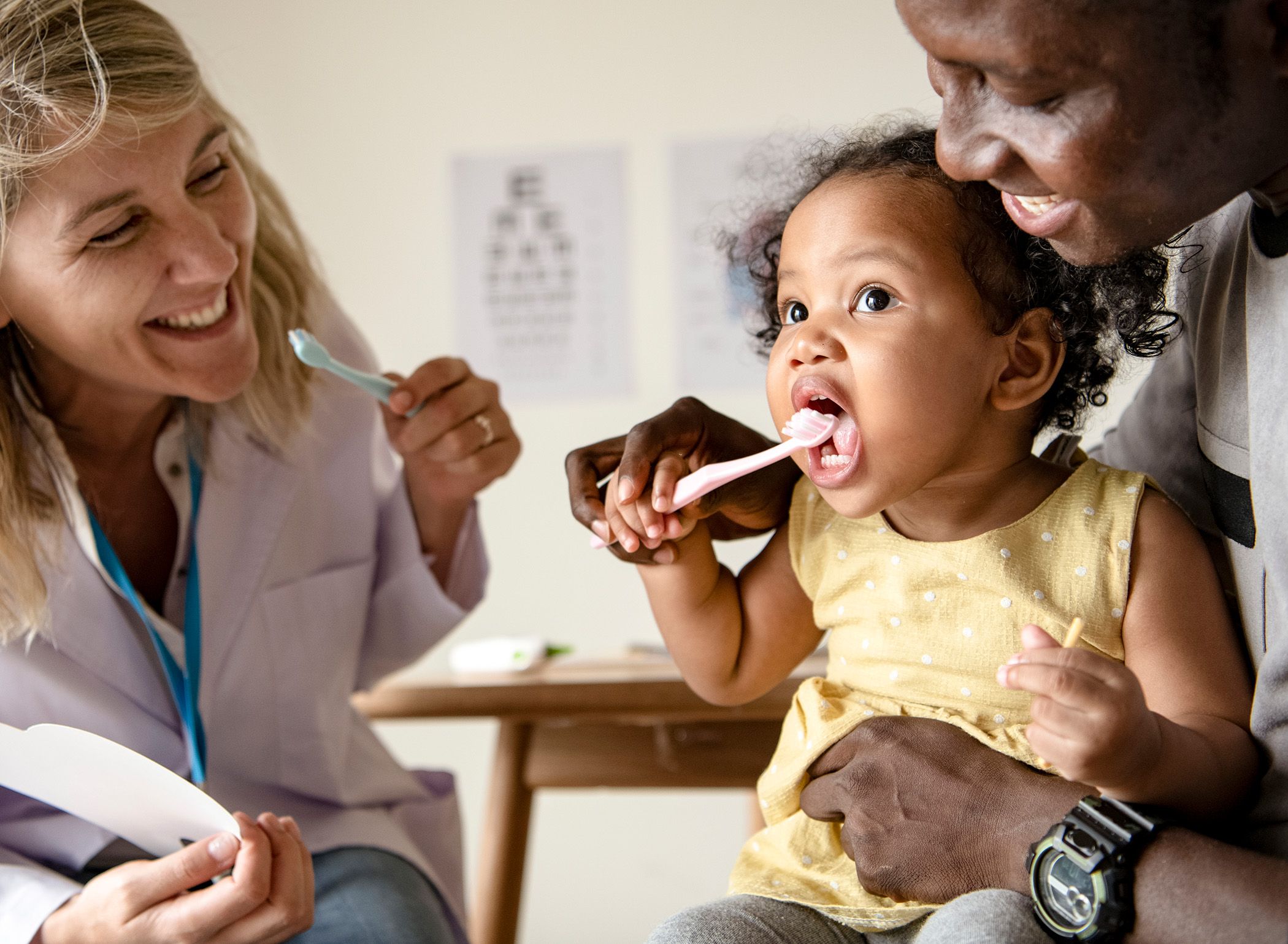 Image of a child being taught how to brush their teeth by her parent and dentist by her side.