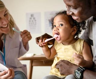 Image of a child being taught how to brush their teeth by her parent and dentist by her side.