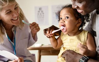 Image of child with her parent showing her how to brush their baby teeth with a dentist present.