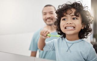Image of a child brushing his teeth with his father in the background.