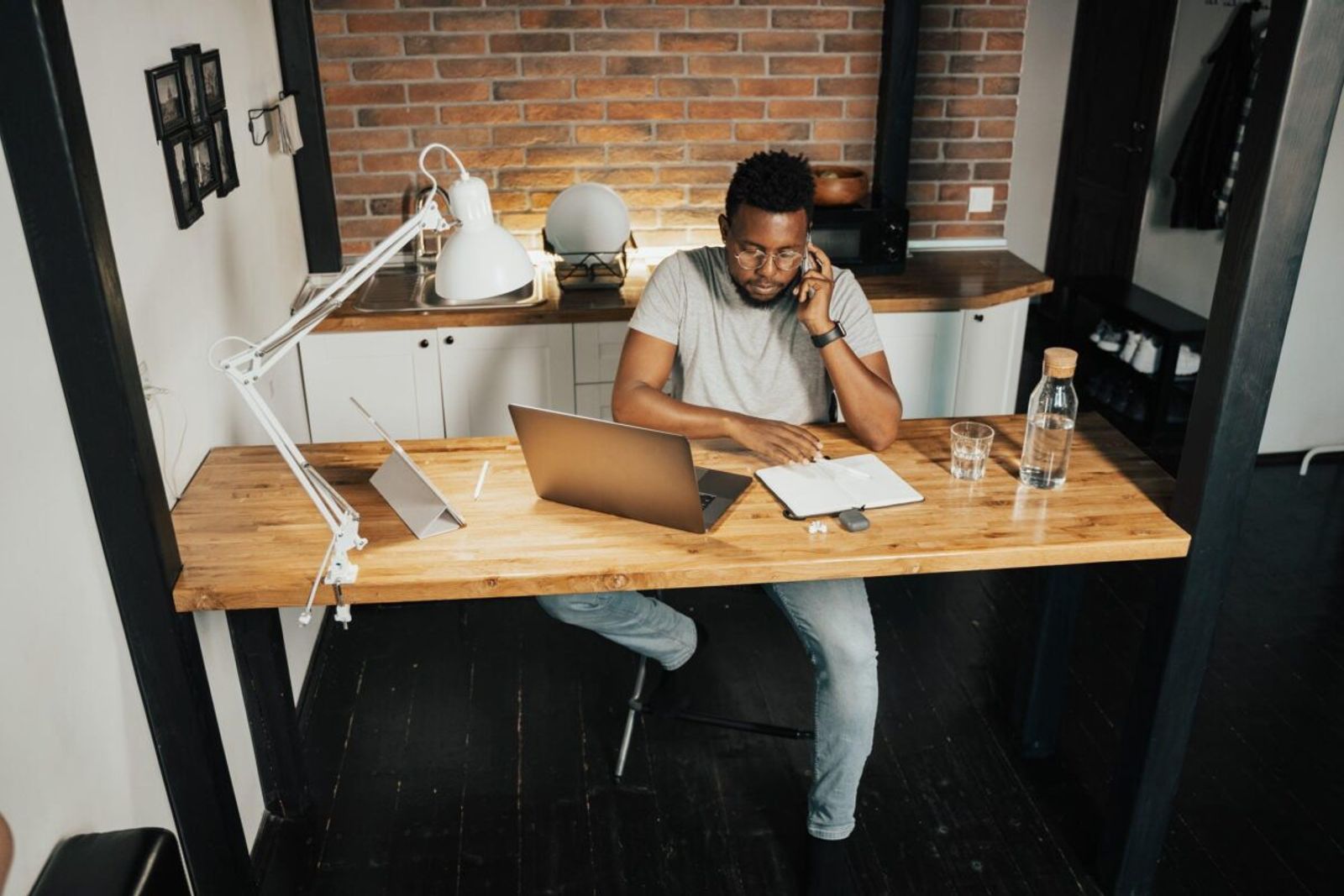 A man on a phone call taking notes beside his laptop at a home desk