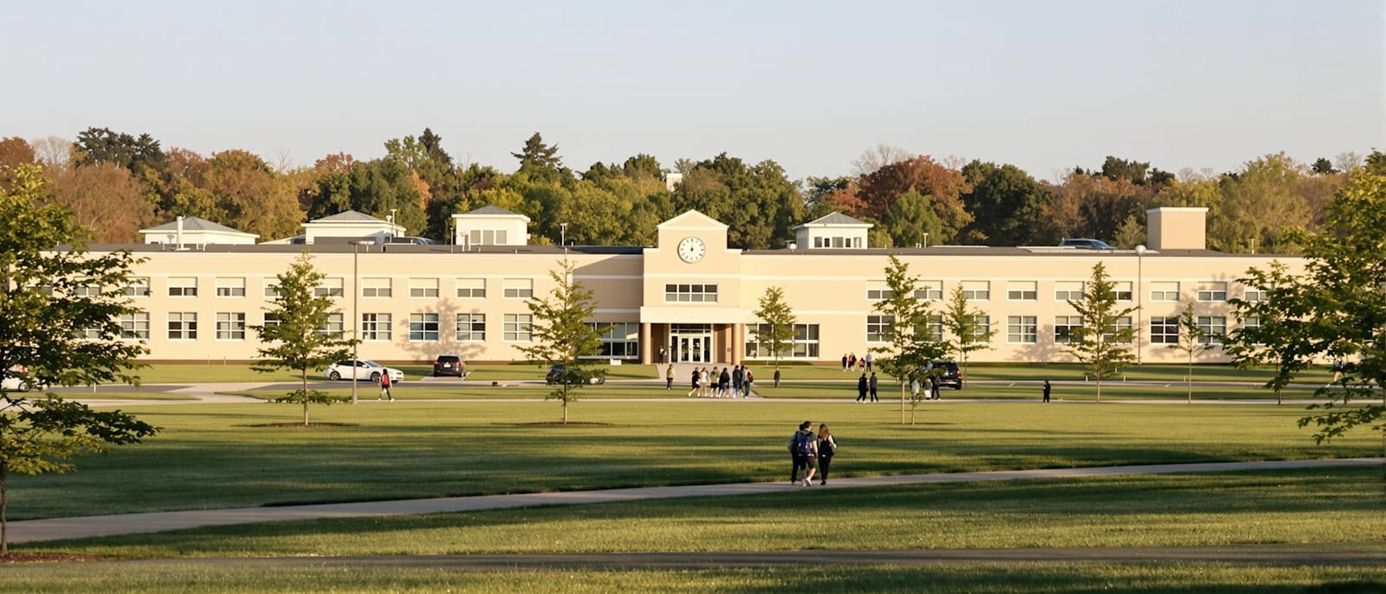 A low suburban school building with students walking across the lawn toward the entrance on a sunny morning.