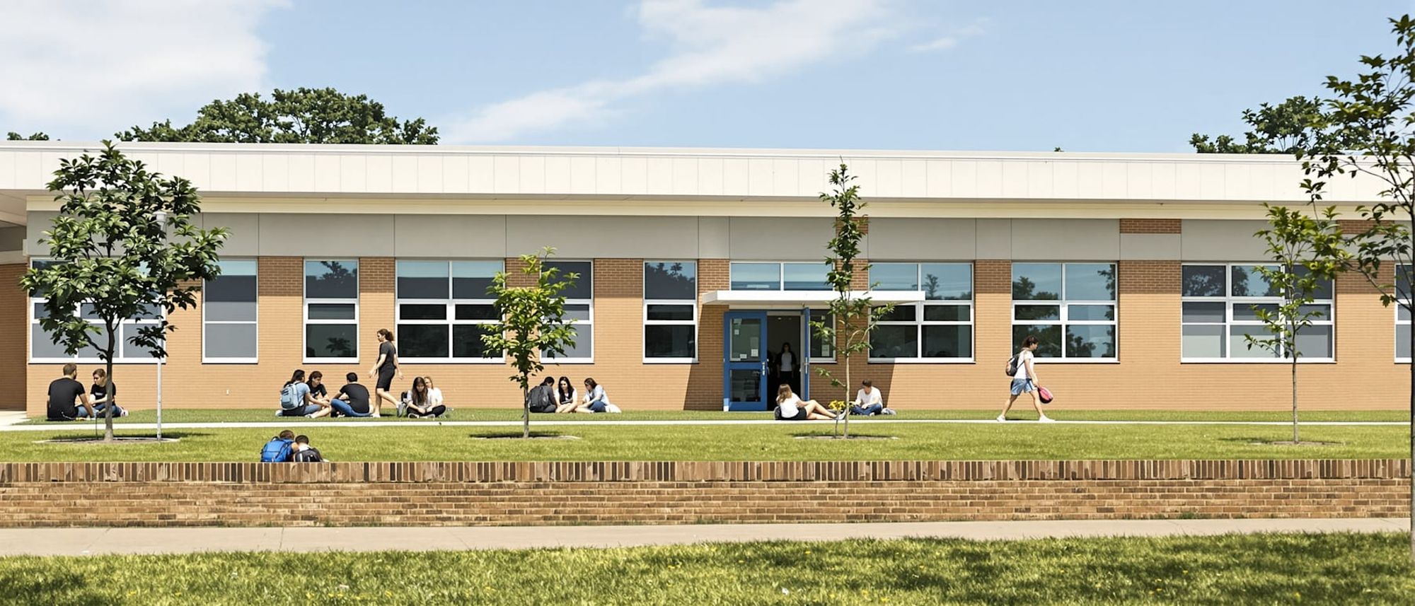 Students sitting and talking on a low wall outside a single-storey school on a sunny afternoon.