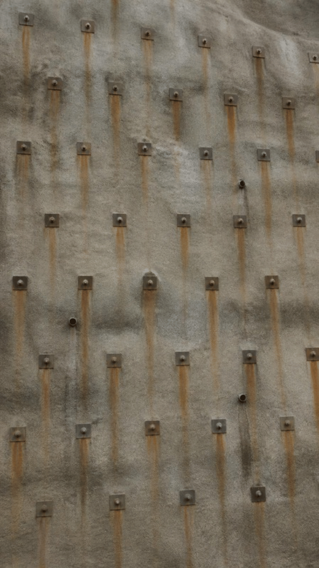 A hillside above a road sprayed with grey shotcrete, with a regular grid of square steel soil nail plates pressed into the surface and rust staining beneath them.