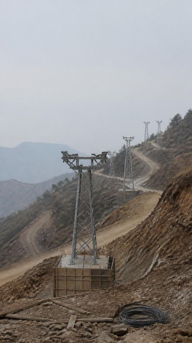Unfinished ropeway tower on a steep hillside with a row of similar towers climbing into the distance.
