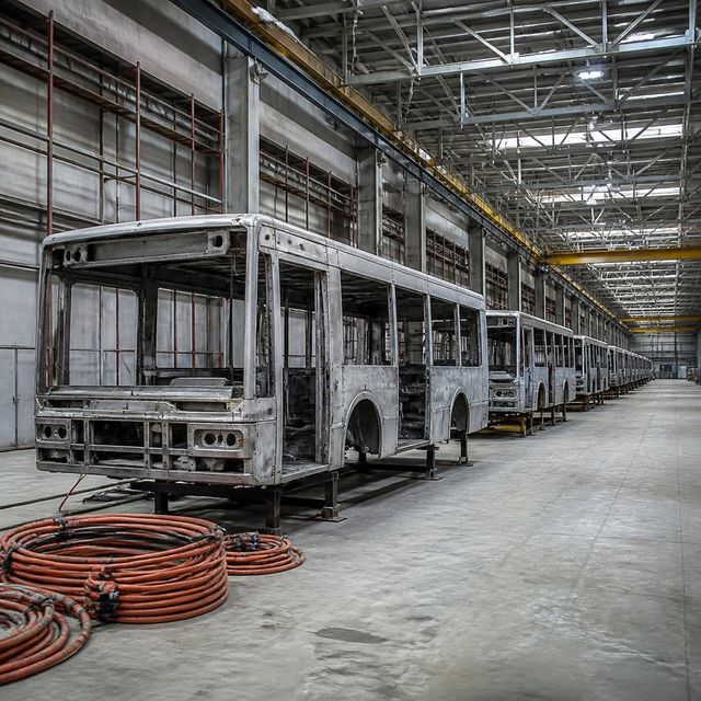 Unfinished electric bus bodies in a row on an assembly line inside a half-built factory hall, receding into the distance.