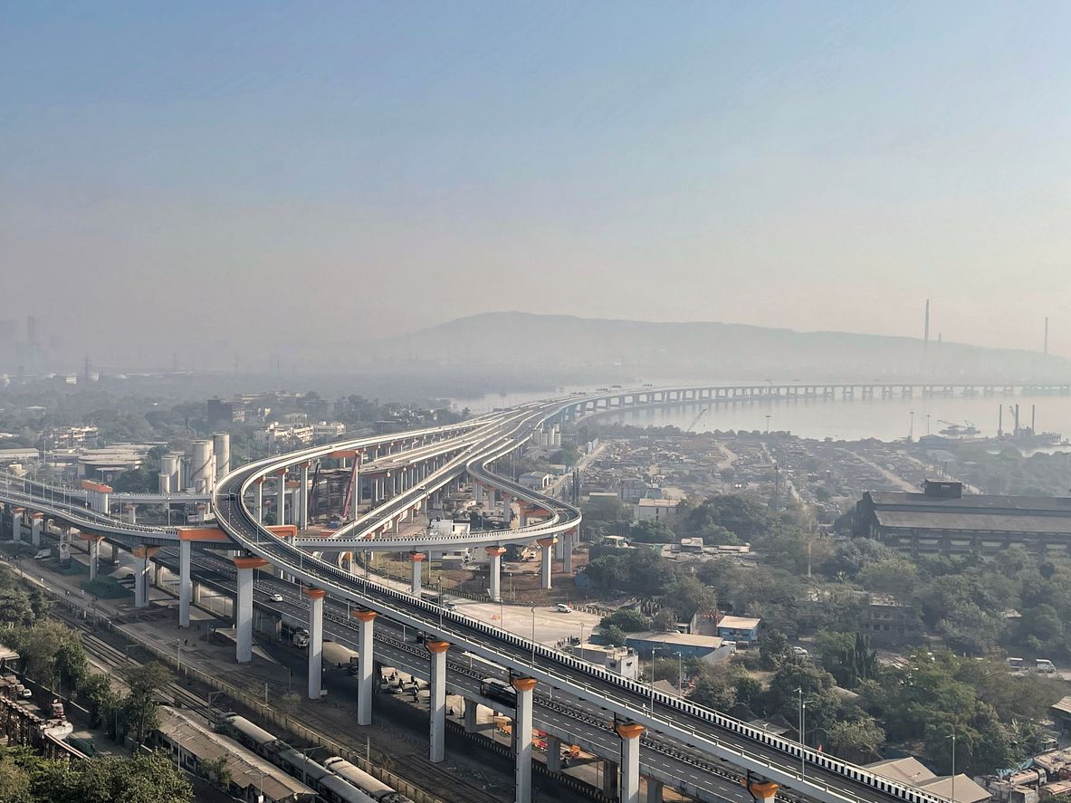 An aerial view of the multi-lane Sewri interchange of the Atal Setu bridge curving over the coastline and urban landscape in Mumbai during a hazy day.