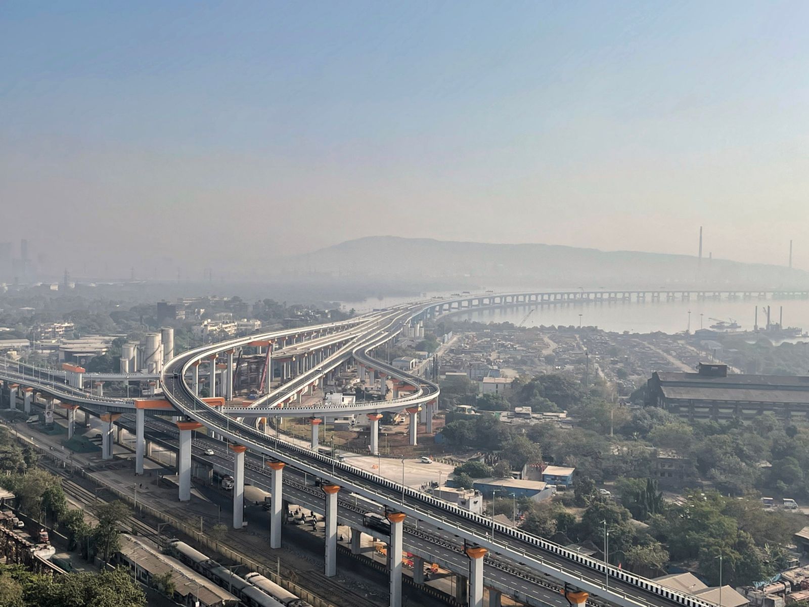An aerial view of the multi-lane Sewri interchange of the Atal Setu bridge curving over the coastline and urban landscape in Mumbai during a hazy day.