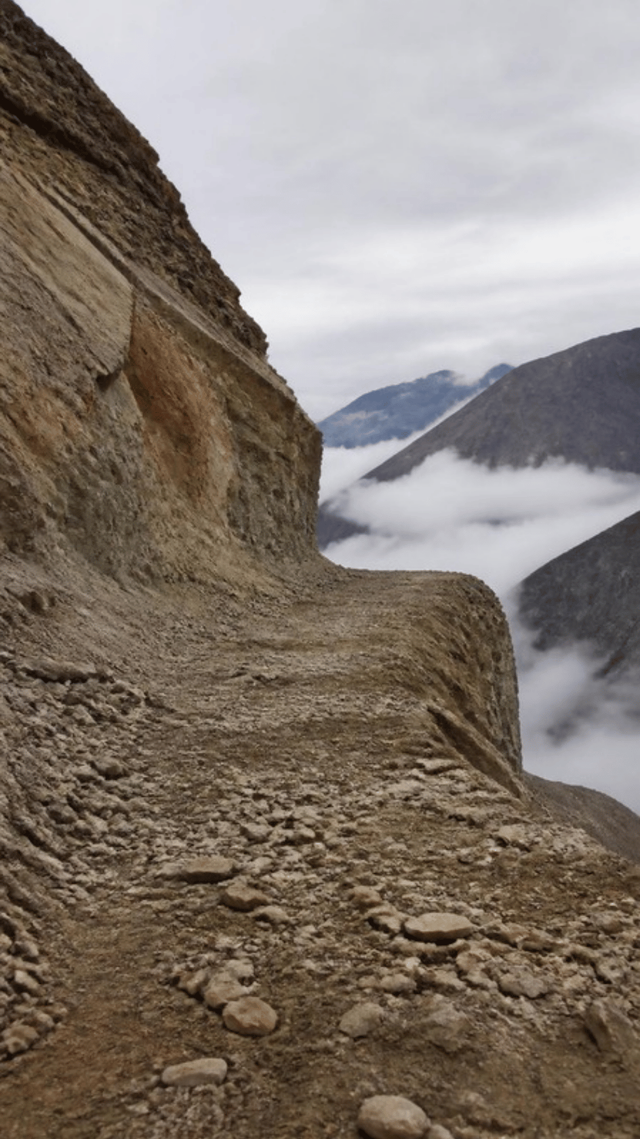 A flat bench cut into a steep Himalayan hillside, ending abruptly at raw rock, with loose material fanned across it and cloud in the valley beyond.