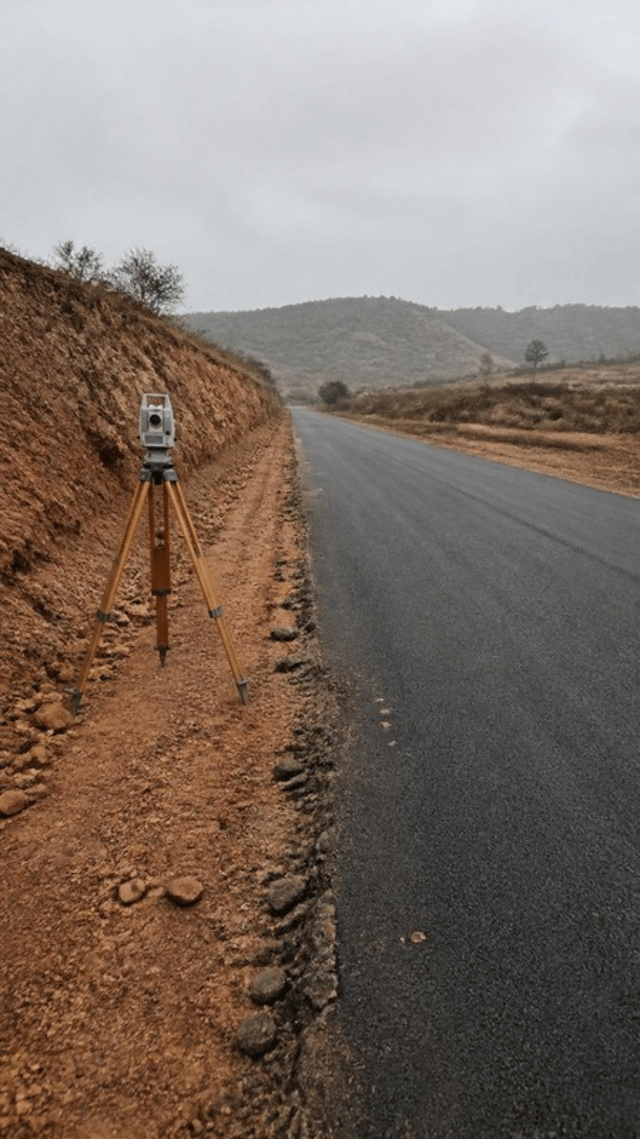 A two lane bitumen road running through a red laterite cutting, with a surveyor's total station on a wooden tripod standing unattended on the earth verge.