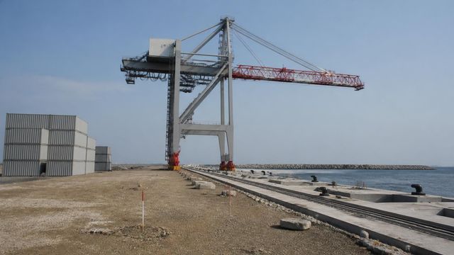 Unfinished container terminal quay with a large crane and a long row of concrete deck segments receding along the water.