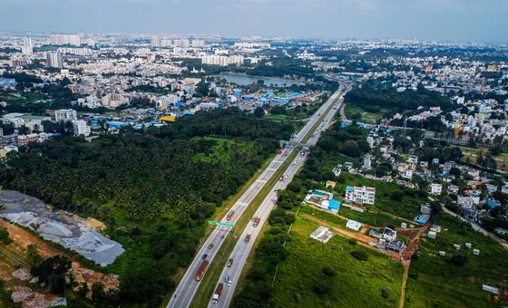 A high-altitude shot looking down the straight, wide Bengaluru-Mysuru Expressway. The highway cuts directly between dense urban housing on the left and open agricultural green spaces on the right.