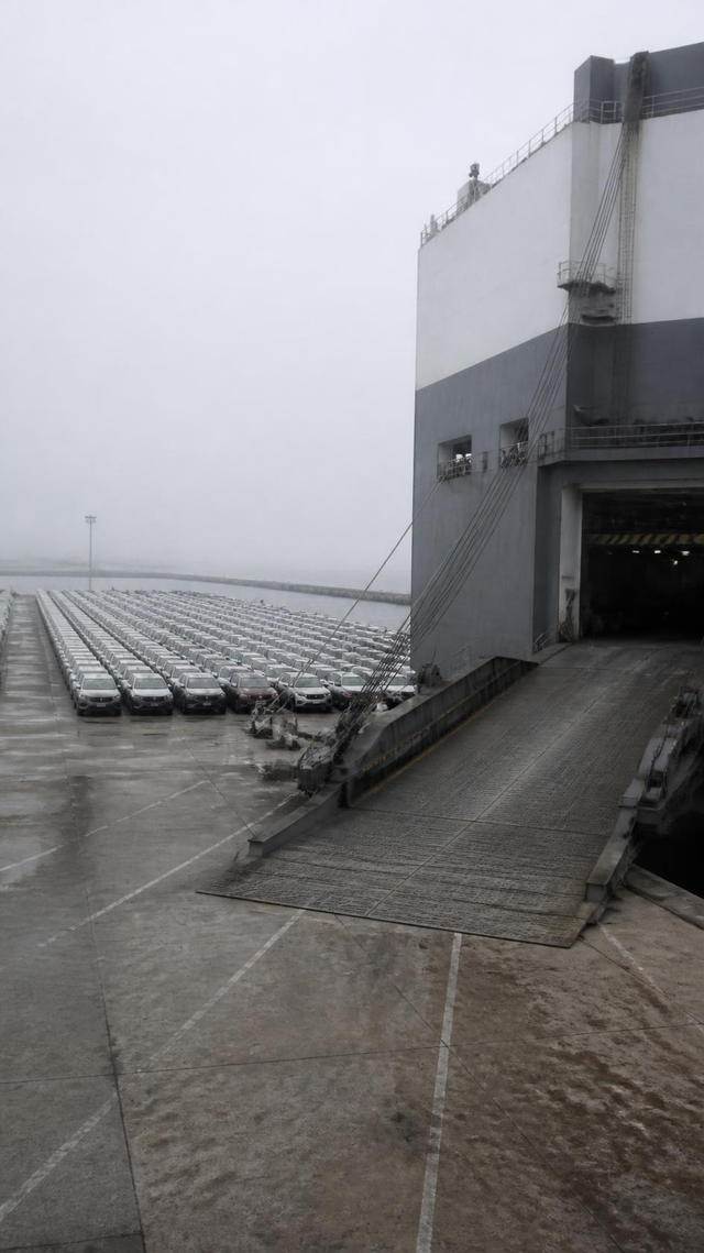 Vehicles staged on a port apron beside the stern ramp of a car carrier, no people present
