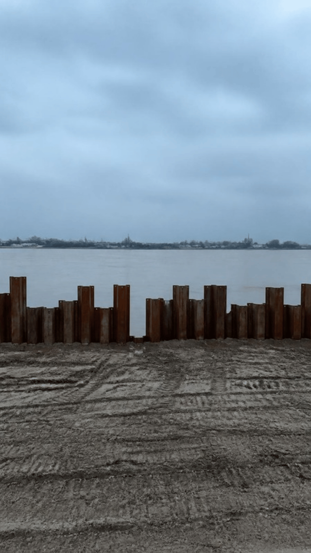 A raw river bank with a ragged line of rust coloured steel sheet piles driven into it at uneven heights, bare levelled earth behind and the wide brown Hooghly beyond.