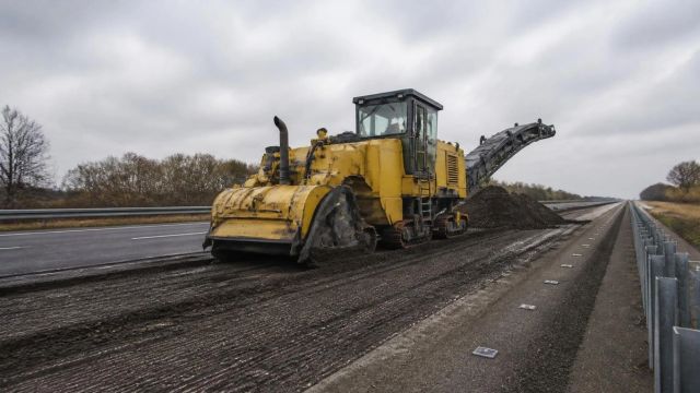 Milling machine on a half-stripped highway lane with crash barrier running to the horizon during road renewal work.