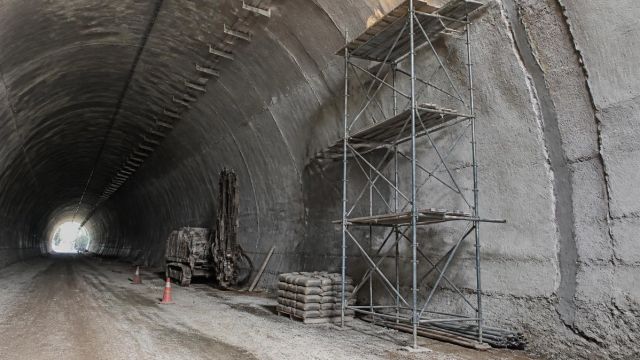 Road tunnel under rehabilitation with scaffold, patched lining and a row of fittings leading to a distant bright exit.