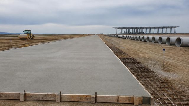 Unfinished concrete taxiway slab with formwork and rebar, receding past edge fixtures toward a bare steel terminal frame.