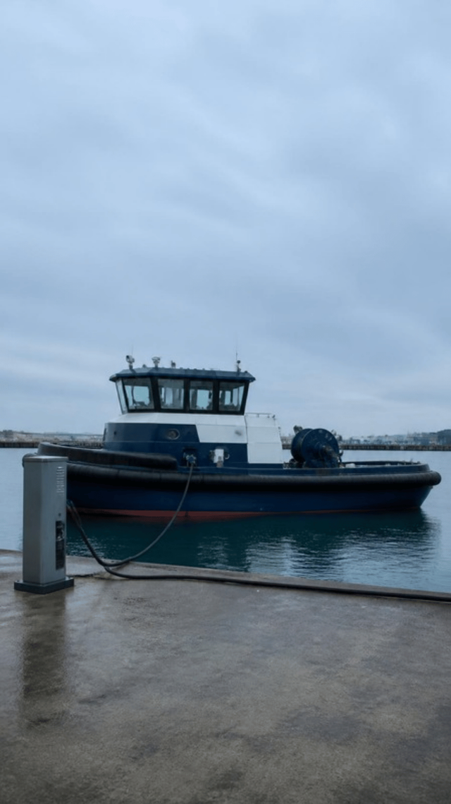 A harbour tug moored alongside a quay with a thick charging cable running from a grey cabinet on the quay to a socket in the hull.